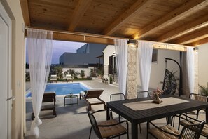 Een terras buiten met een zwembad, houten plafond, witte gordijnen en een eethoek met stoelen en een tafel.