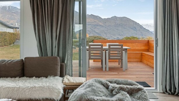 Una habitación con un amplio ventanal corredera que da a una terraza exterior con mesa y sillas de madera, con vistas a un paisaje montañoso.