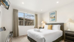A hotel room with a large bed, a desk, a chair, a window with curtains, a nightstand with a lamp, and a painting on the wall. - Amazing Location! Central 2 bedroom Unit (Wānaka)