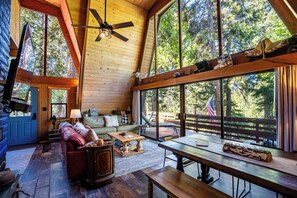 Interior acogedor de una cabaña con techo de madera, ventanas amplias, sofá, mesa de centro y mesa de comedor.