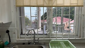 A kitchen with a sink, faucet, and a window with a view of buildings and a red roof. - Tropical Paradise Retreat in Soufriere (Soufriere)