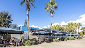 Terraza al aire libre con sombrillas, palmeras y un edificio con letrero 'CAFFE NERO'.