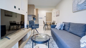 Una sala moderna con un sofá, una mesa de centro y sillas. Hay un comedor con una mesa y sillas. La habitación tiene armarios de madera y una cocina al fondo.