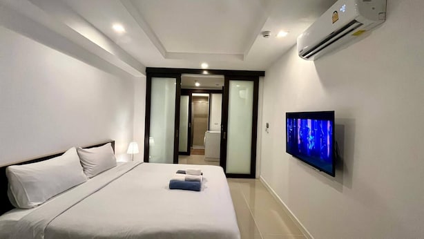 A hotel room with a bed, a flat-screen TV mounted on the wall, an air conditioning unit, and a door leading to a bathroom. - 6-15 - 1 Bdr Condo With Balcony and Shared Pool (Patong)