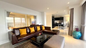 A modern living room with a dark brown sofa, a glass coffee table, and a beige ottoman. There is a dining area with a glass table and chairs, and a kitchen visible in the background. - A11 - 2 Bdr Condo, Stunning Sea View, Karon Beach (Karon)