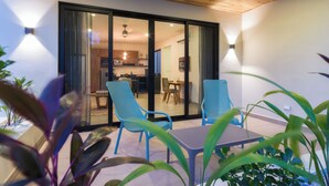 Un patio moderno con dos sillas azules y una mesita pequeña, con vistas a un comedor interior con muebles de madera y una cocina visible a través de puertas de cristal.