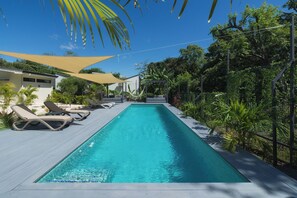 Une piscine rectangulaire, dotée de chaises longues et d’un espace ombragé, entourée d’une végétation luxuriante et surplombée d’un ciel bleu azur.