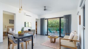Un salon moderne comprenant une table à manger, des chaises, un canapé et une petite table basse. Une grande fenêtre, ornée de rideaux, offre une vue sur la verdure environnante.