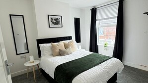 A modern bedroom with a black headboard, a green throw blanket, and a white bedspread. There is a small bedside table with a lamp, a framed picture on the wall, and a window with black curtains.