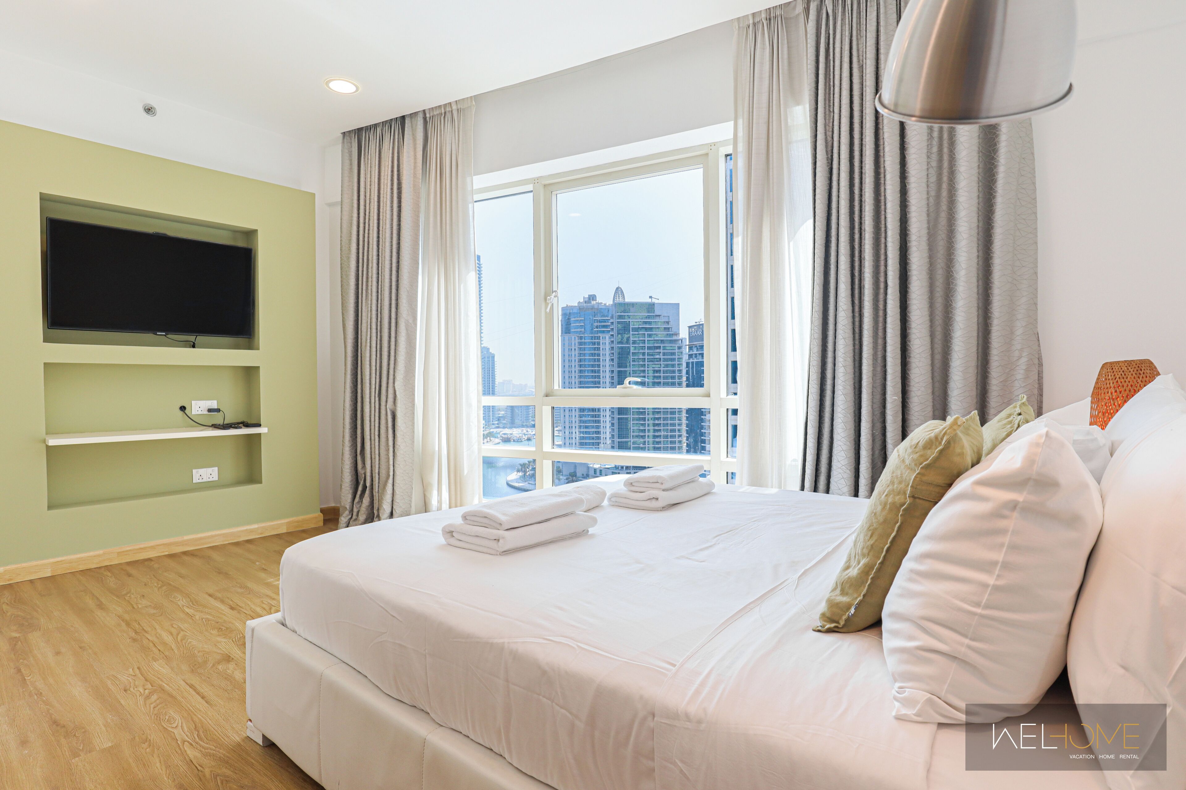 A modern hotel room with a large bed, a flat-screen TV mounted on the wall, and a view of the cityscape through the window.