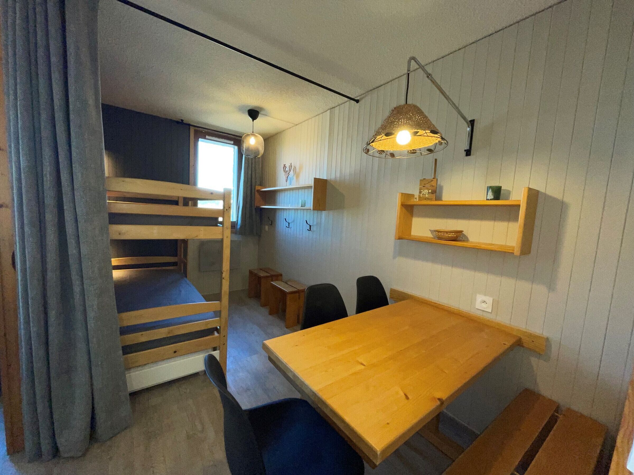 A room with bunk beds, a wooden table, chairs, a hanging light fixture, and wall-mounted shelves.