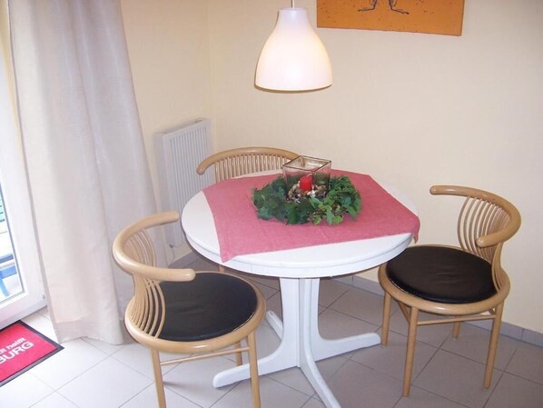 Un pequeño comedor con una mesa blanca, dos sillas de madera con cojines negros, un mantel rojo sobre la mesa y una lámpara colgante arriba.