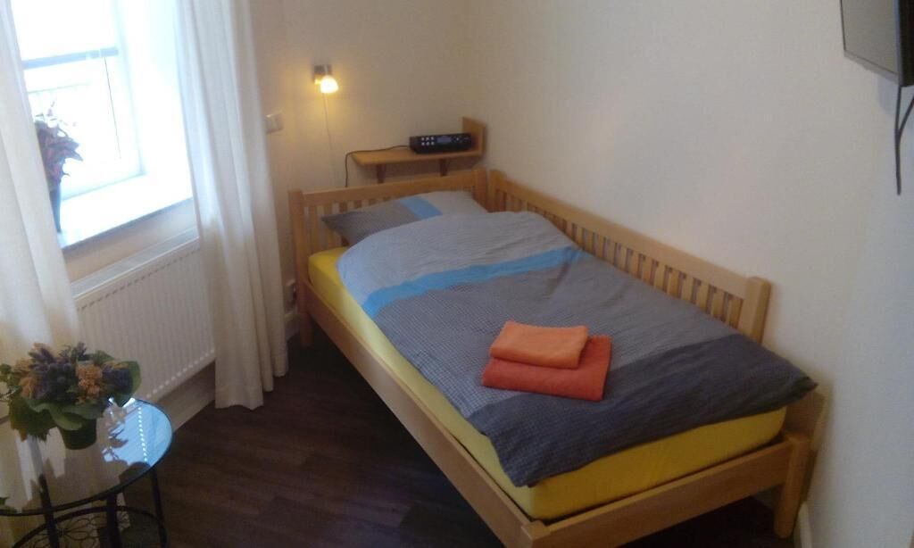 A small, single-bed room with a wooden frame, a bedside table with a radio, a window with curtains, and a small table with a plant.