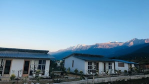 A row of white buildings with metal roofs, surrounded by a fence and potted plants, with a mountain range in the background.