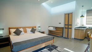 A modern bedroom with a large bed, bedside tables, a built-in bench, a small kitchenette, and a door leading to a bathroom.