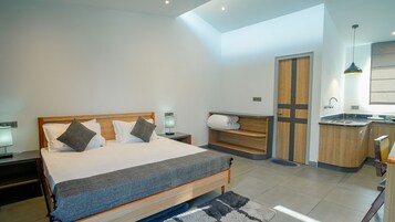 A modern bedroom with a large bed, bedside tables, a built-in bench, a small kitchenette, and a door leading to a bathroom.