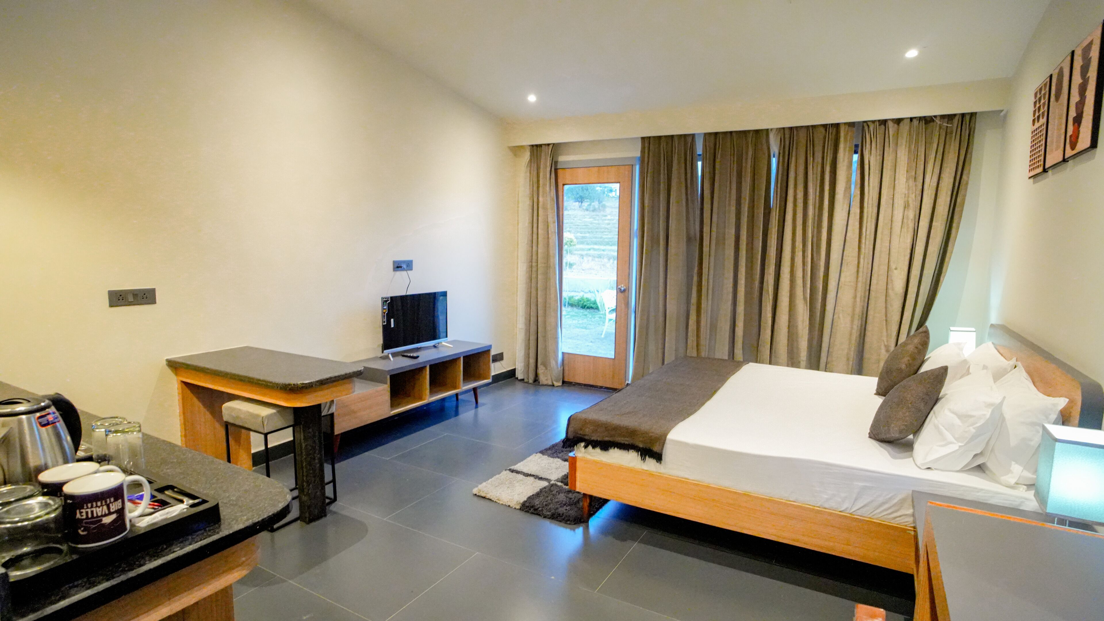A hotel room with a bed, a desk with a television, a coffee station, and a view of the outdoors through a door.