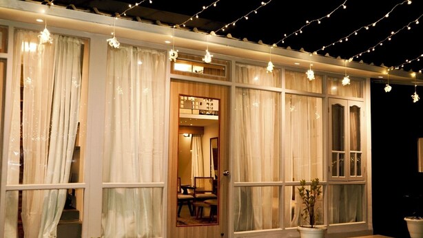 A hotel room with large windows, a wooden door, and string lights. There are potted plants on the windowsill.