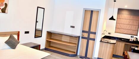 A modern hotel room with a bed, a small desk, a mirror, a wardrobe, and a kitchenette.