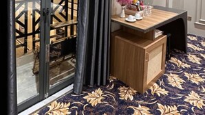 A hotel room with a sliding glass door, a flat-screen TV mounted on the wall, a small wooden desk with a vase of flowers, and a patterned carpet. - Phuoc Hung 3 (Long Chau)