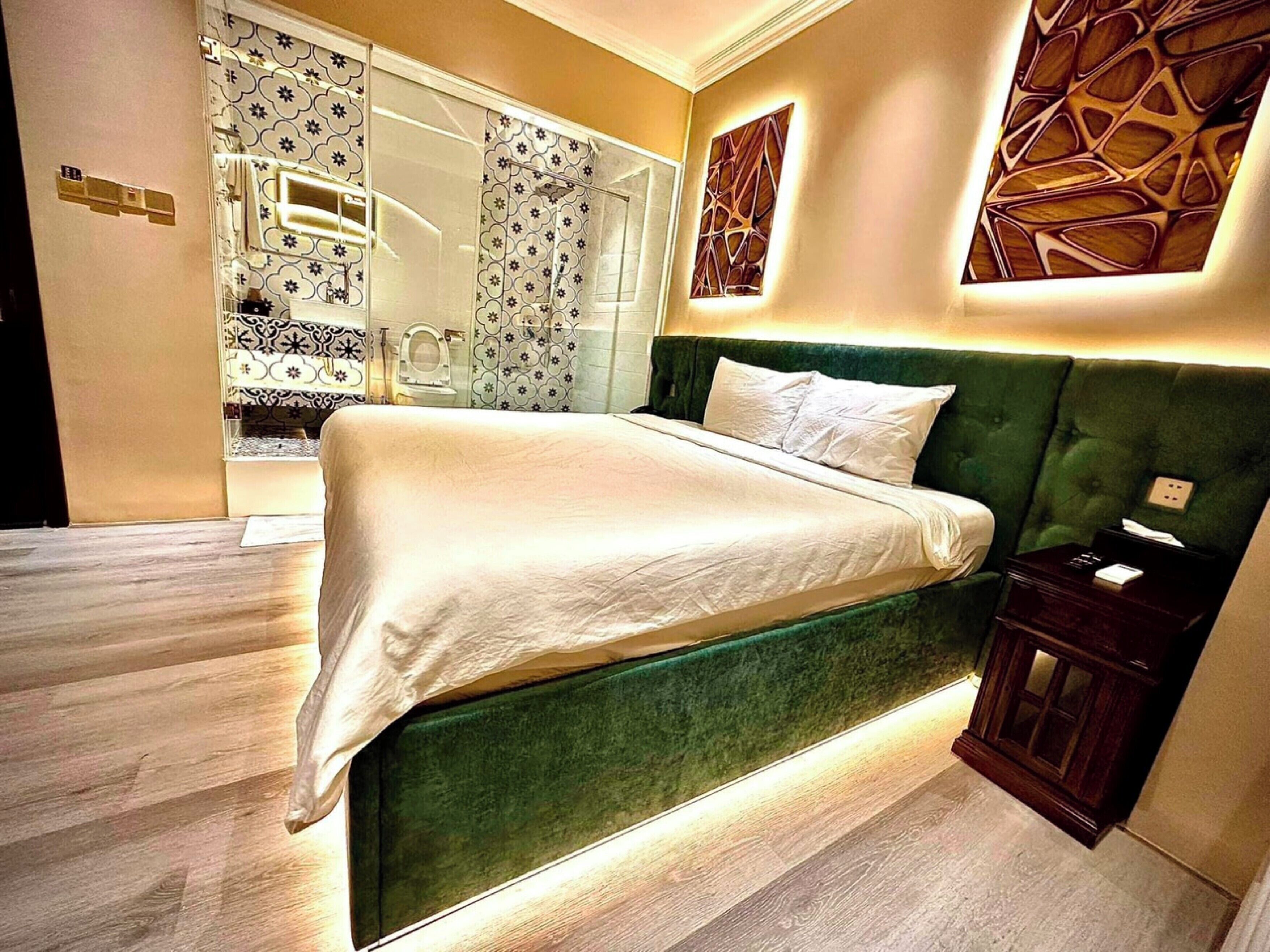 A modern bedroom with a green upholstered headboard, a large bed with white linens, a wooden nightstand, and a decorative wall panel.