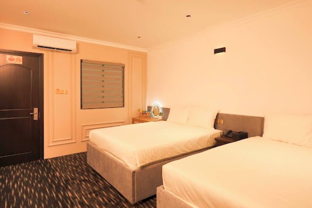 A hotel room with two single beds, a dark door, an air conditioning unit, a window with blinds, a bedside table with a lamp, and a patterned carpet.