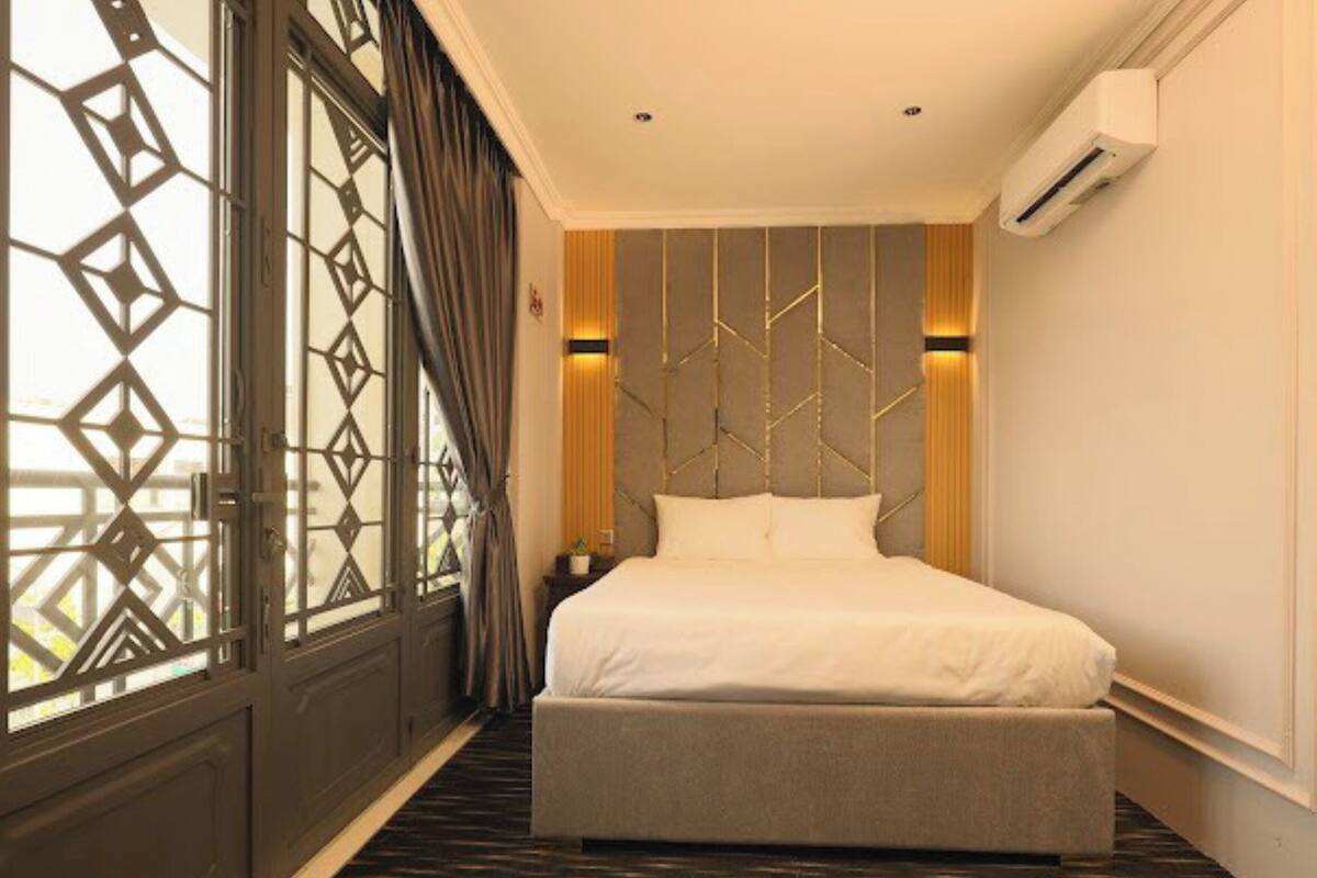 A hotel room with a large bed, a nightstand, a wall-mounted air conditioner, and a window with a geometric pattern.