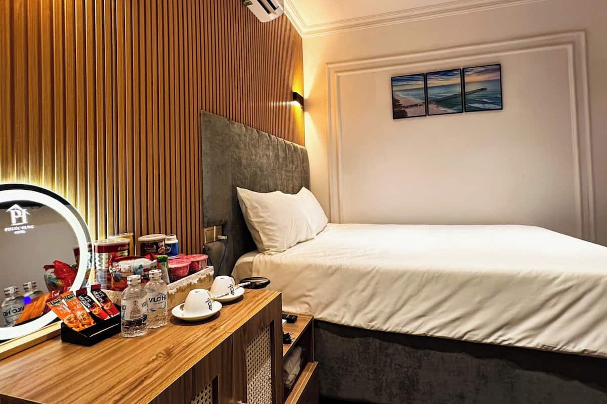 A hotel room with a wooden panel wall, a bed with white bedding, a bedside table with toiletries, and a painting on the wall.