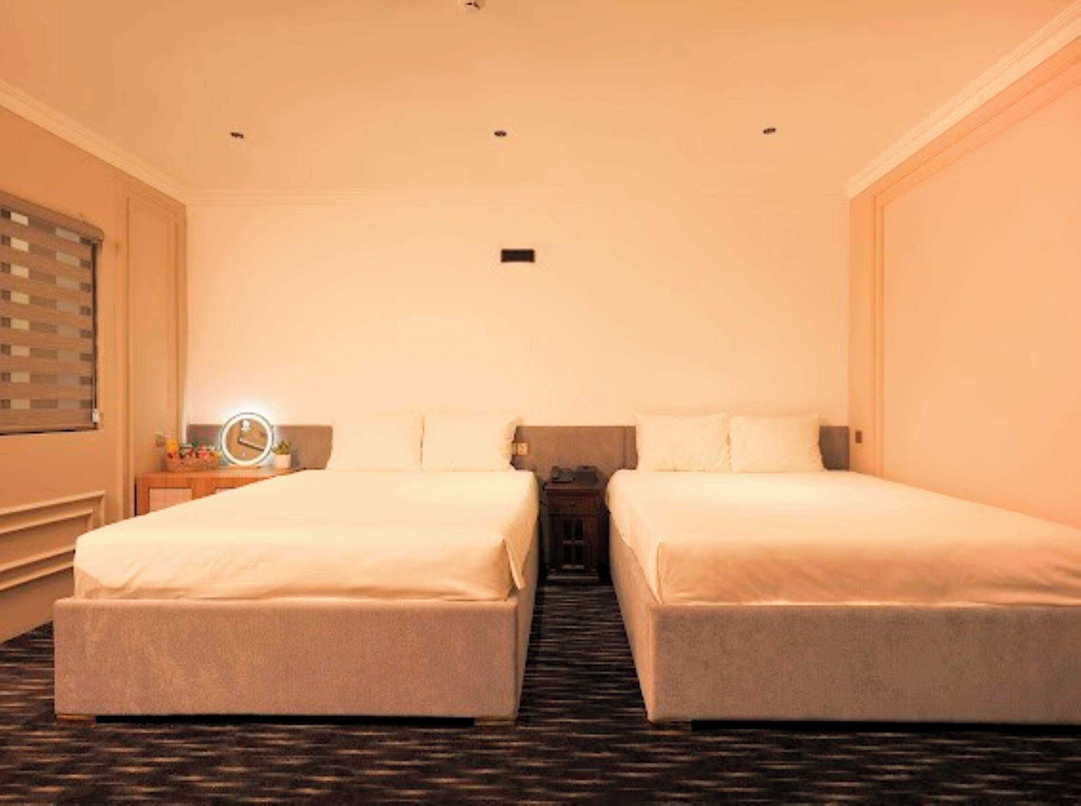 Ein Hotelzimmer mit zwei Einzelbetten, einem kleinen Nachttisch mit Lampe, einer Uhr und einem Spiegel.