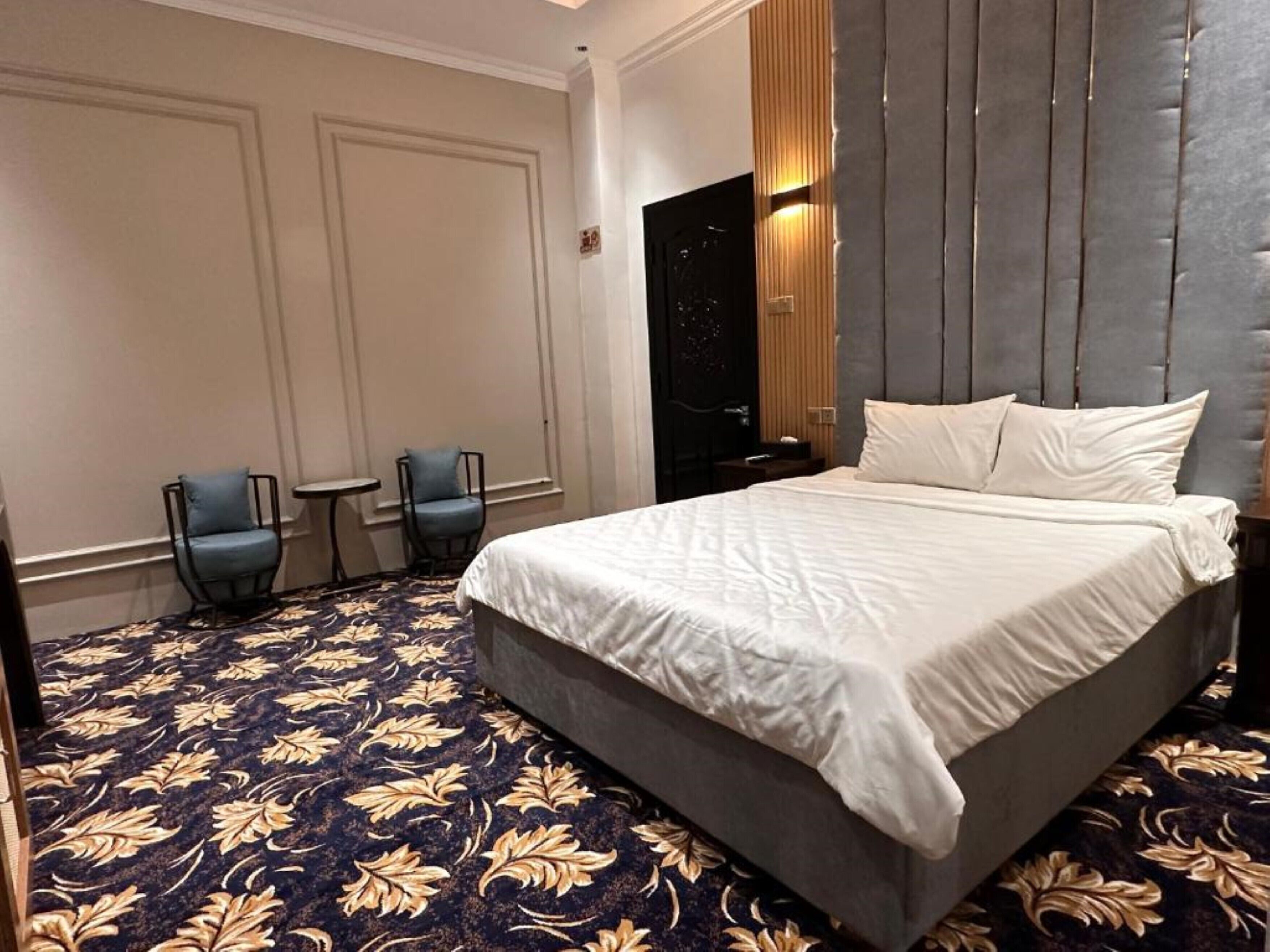 A hotel room with a large bed, two chairs, a small table, and a dark patterned carpet.