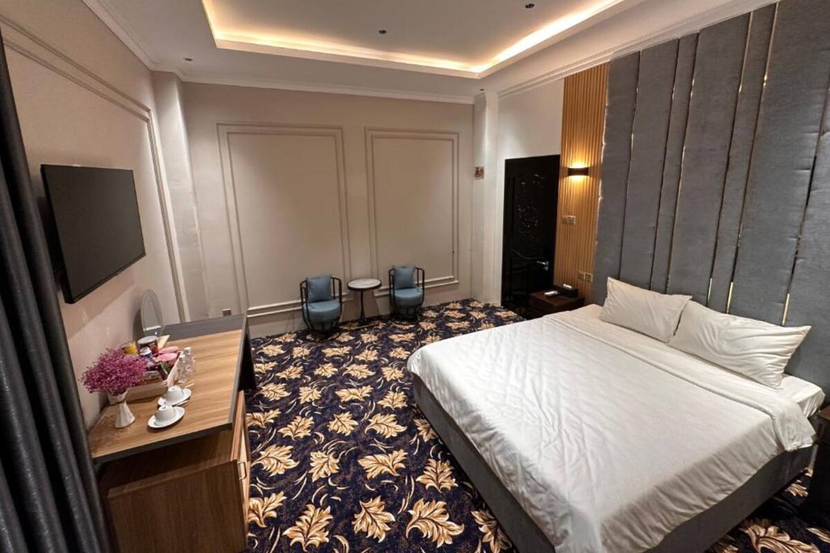 A hotel room with a large bed, a desk with a flat-screen TV, two chairs, and a patterned carpet.
