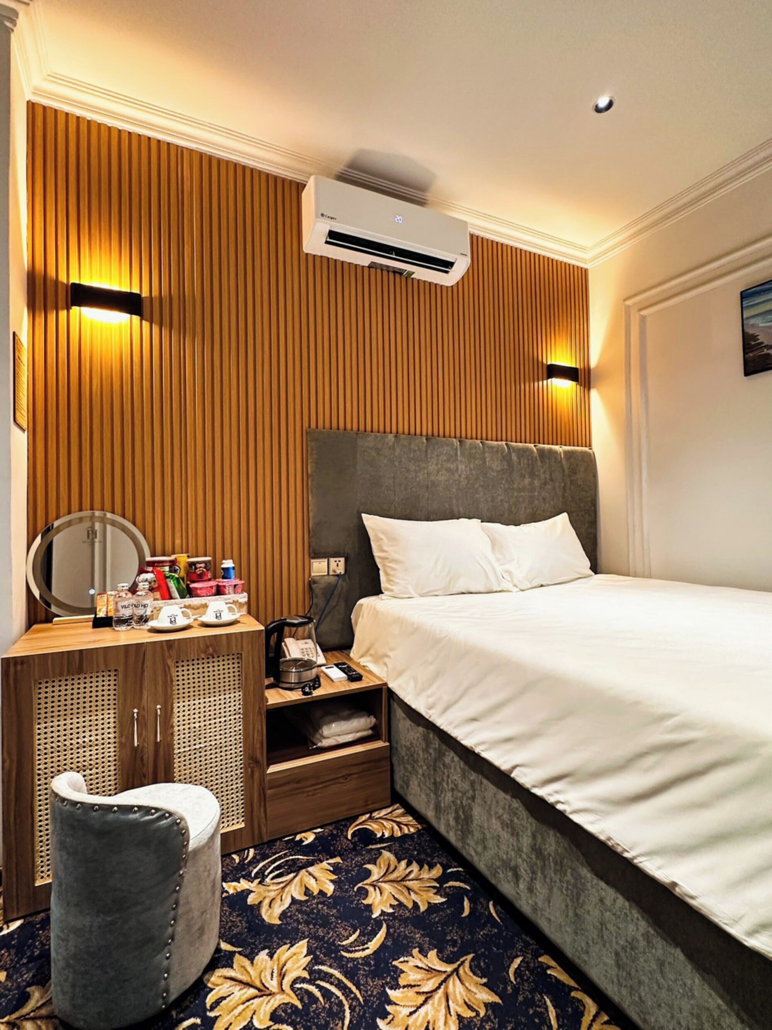 A hotel room with a bed, a wooden wardrobe, a vanity with a mirror, and an air conditioning unit.