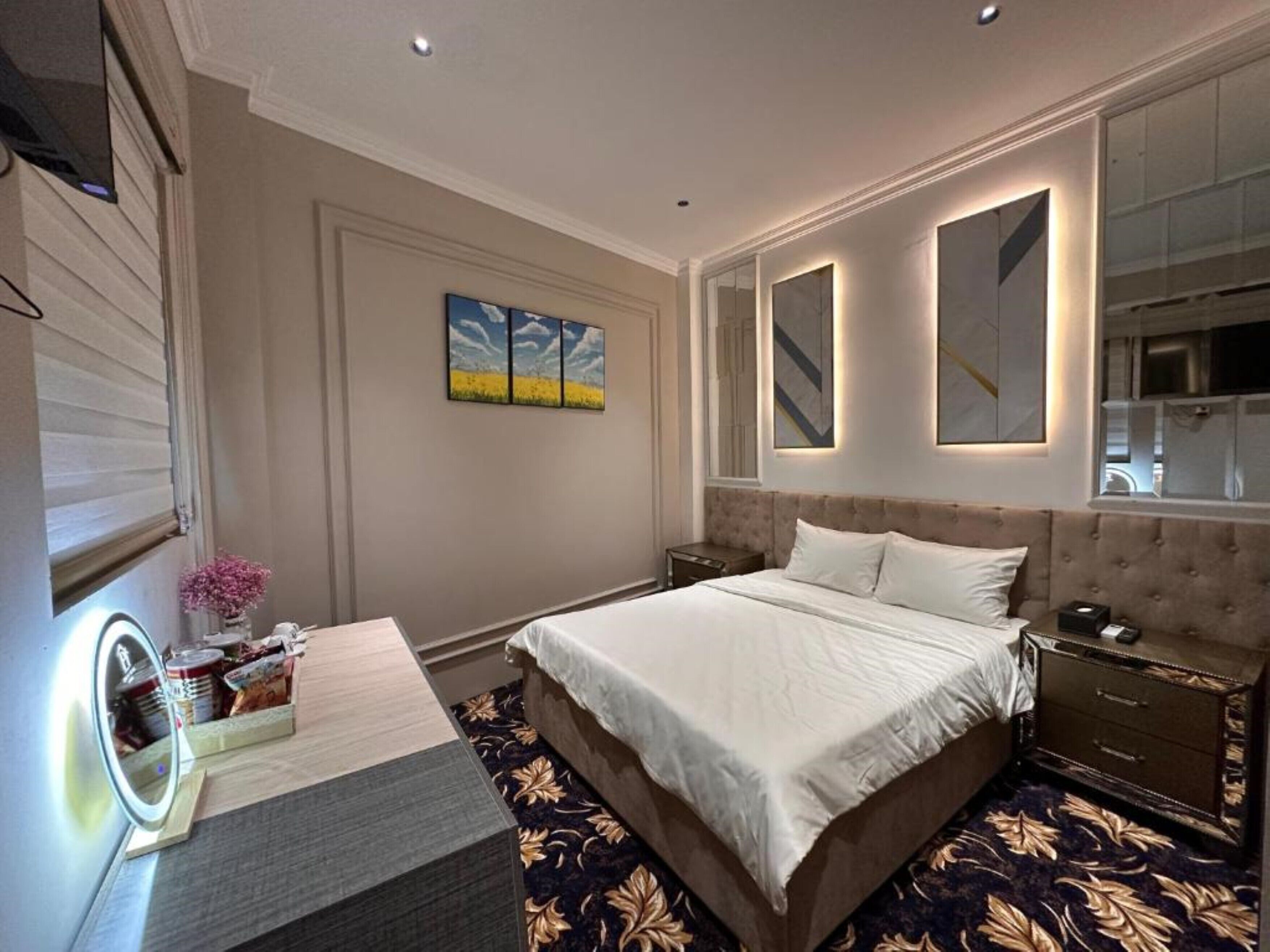 A bedroom with a large bed, bedside tables, a vanity with a mirror, and artwork on the wall.