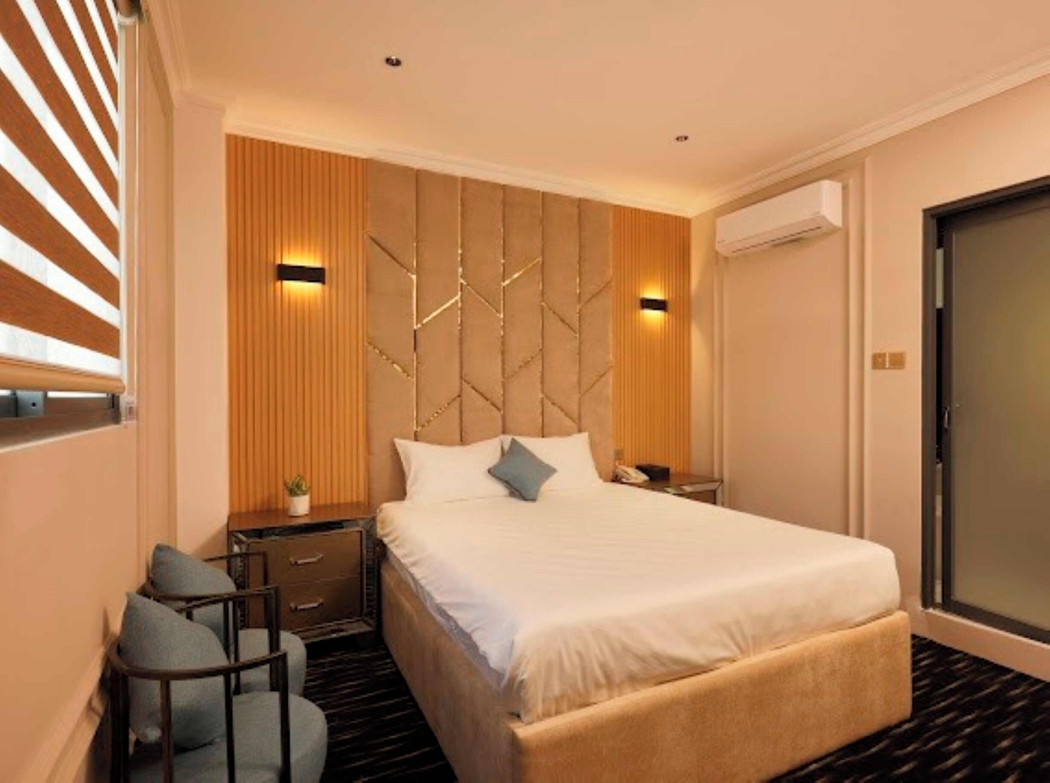 A modern hotel room with a large bed, a desk with a chair, a small plant, and a wall-mounted air conditioning unit.