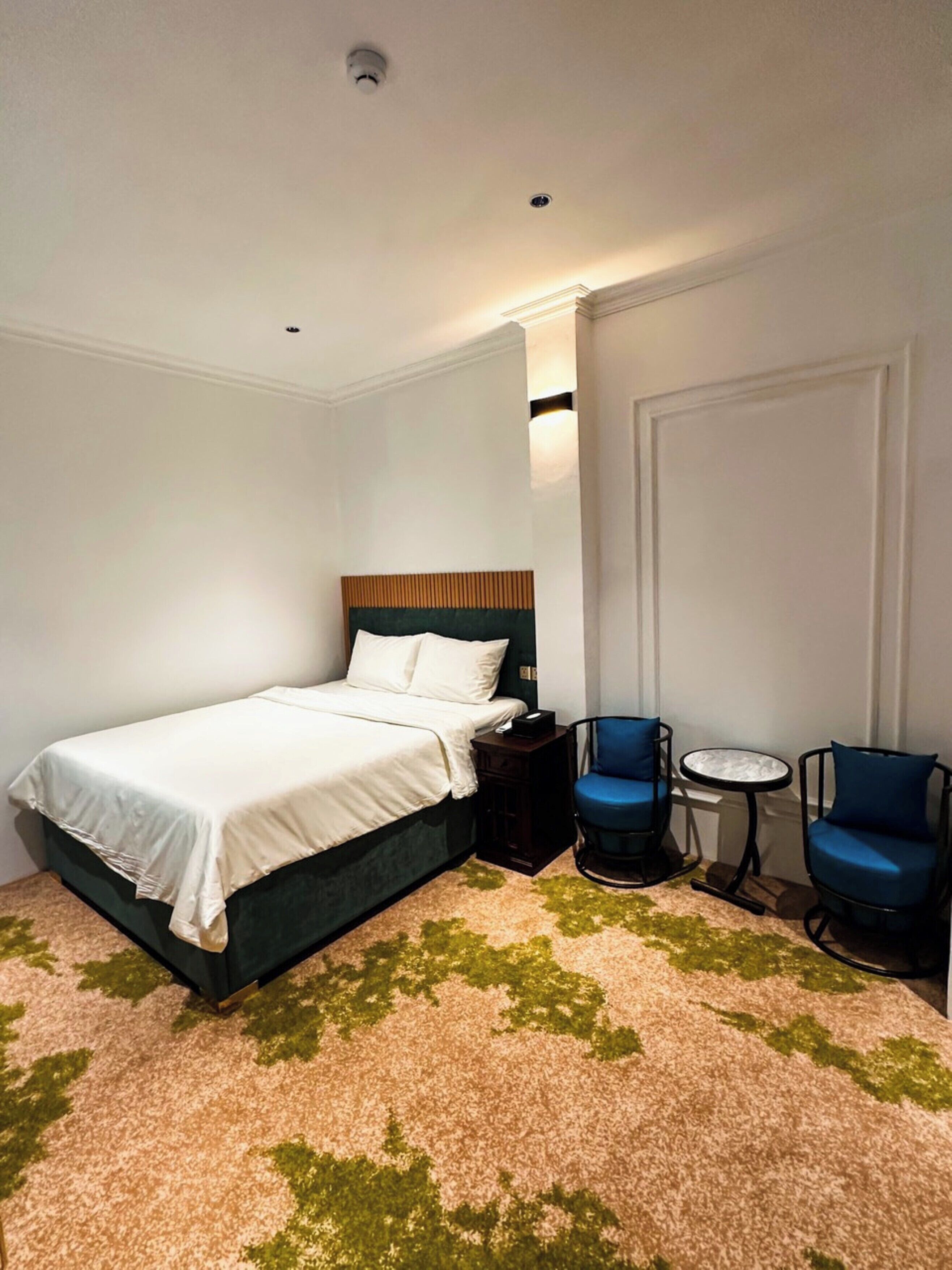 Une chambre d’hôtel avec un lit, deux chaises bleues, une petite table ronde et un tapis à motifs verts.