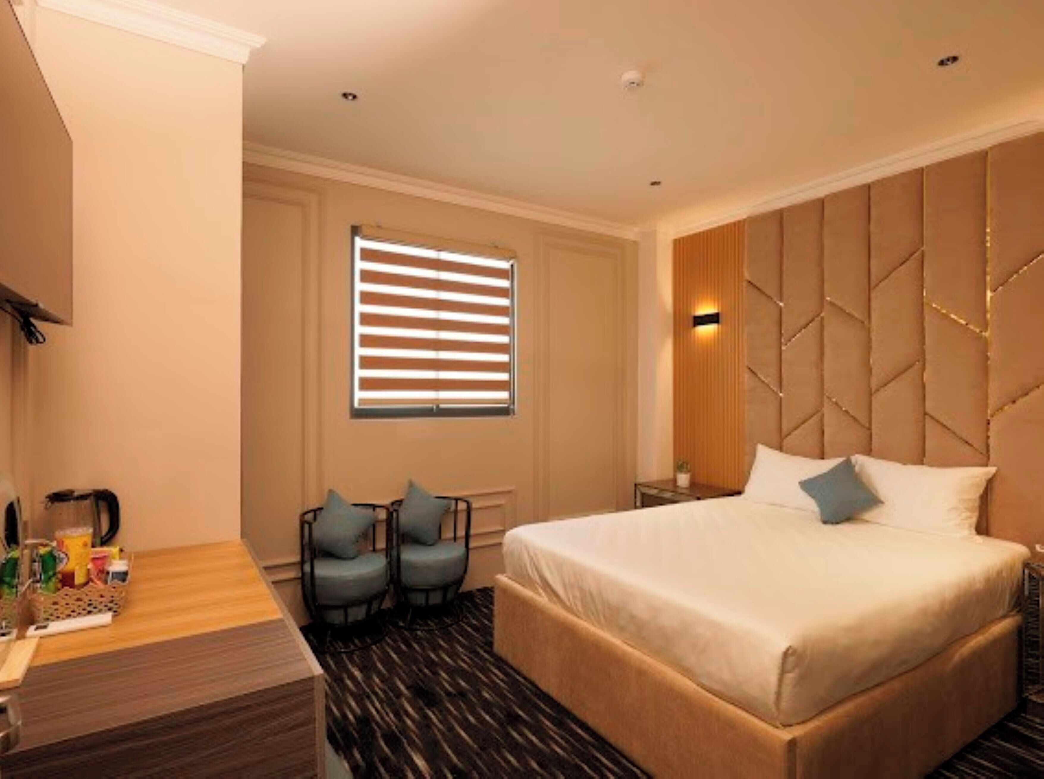 Une chambre d’hôtel avec un grand lit, deux fauteuils, un petit bureau avec une lampe et une fenêtre avec des stores.