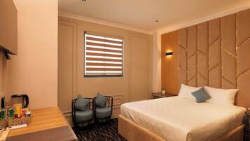 Une chambre d’hôtel avec un grand lit, deux fauteuils, un petit bureau avec une lampe et une fenêtre avec des stores.