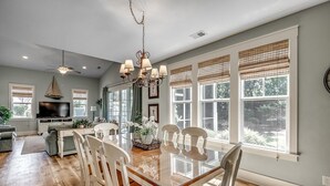 A spacious dining area with a large wooden table and white chairs, a chandelier, and large windows with blinds.