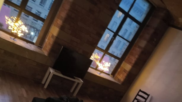 A room with a wooden floor, a black sofa, a small table, and a window with a view of buildings. - 1 Bed Flat - Sleeps 2 - Parking - Wifi (Bradford)