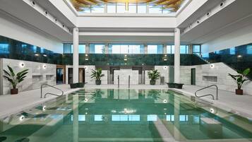 Una amplia piscina interior con base de vidrio, rodeada de paredes blancas y plantas en macetas.