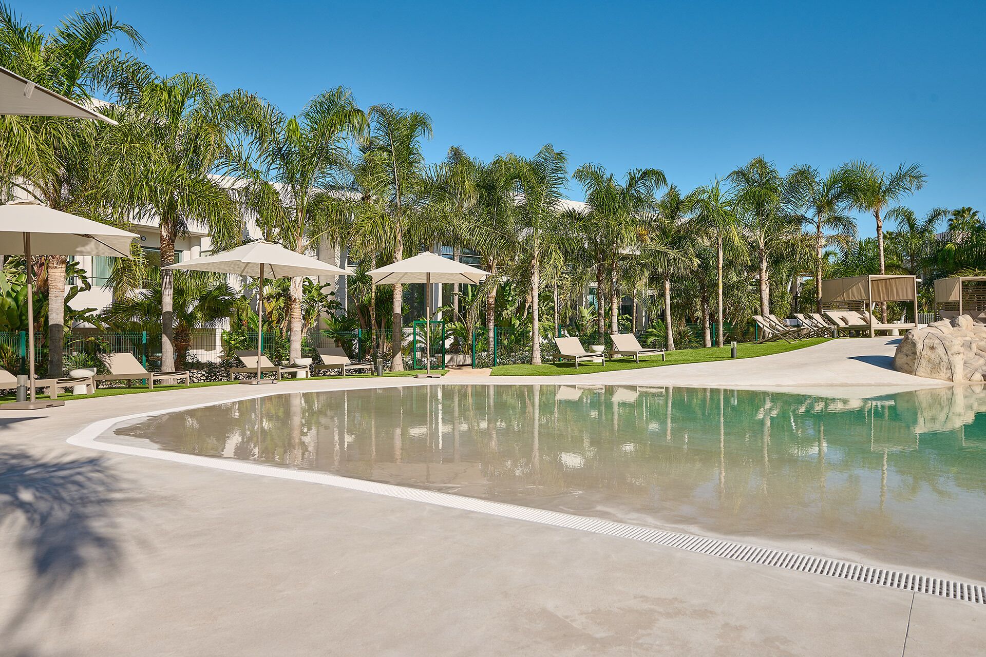 Un área de piscina con sillones de descanso y sombrillas, rodeada de palmeras.