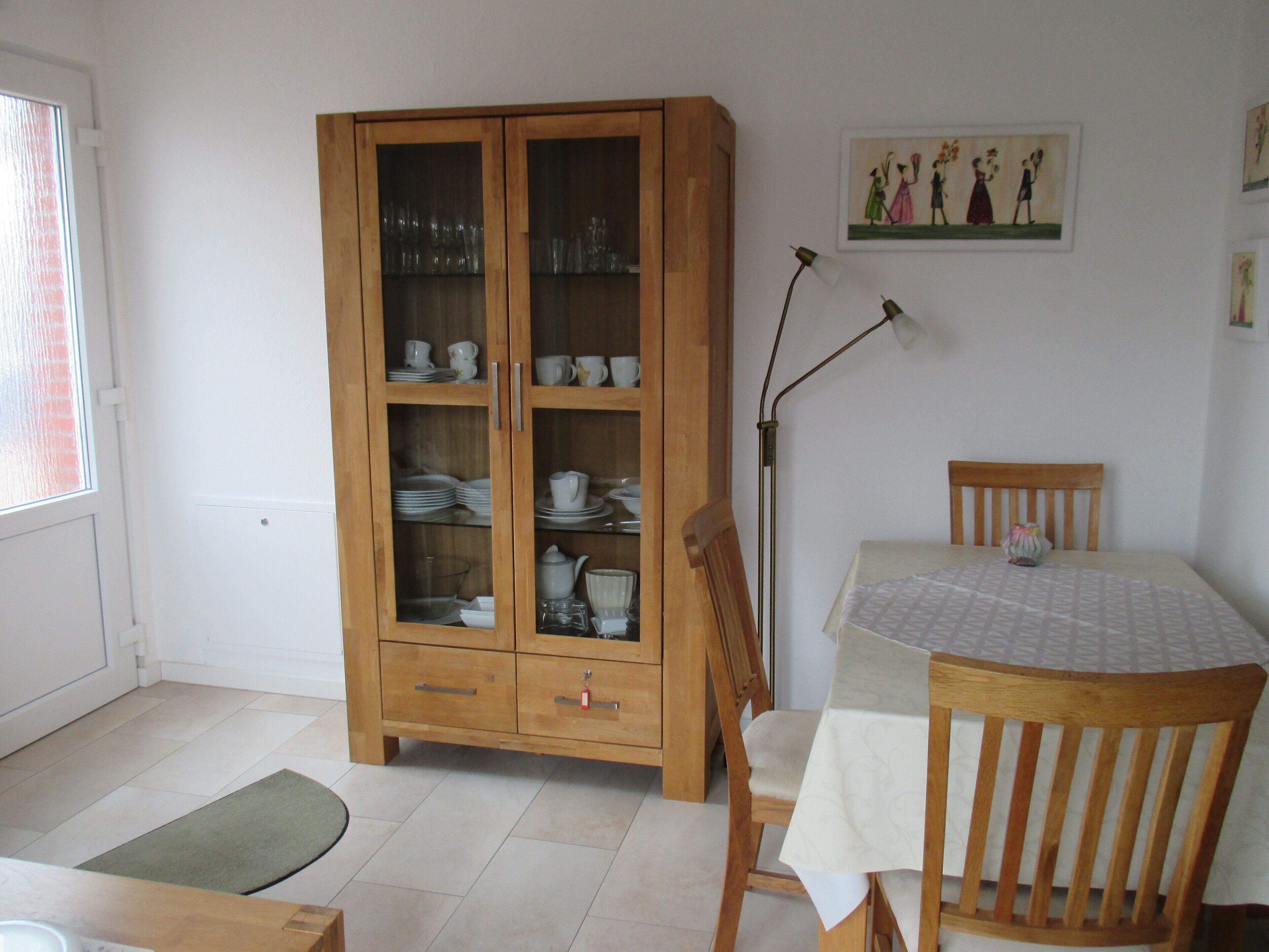 A dining area with a wooden cabinet containing dishes, a table with a tablecloth, and a chair.