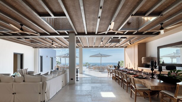 A spacious living area with a large sofa, dining table, and chairs. There are ceiling fans and a view of the ocean through large windows.