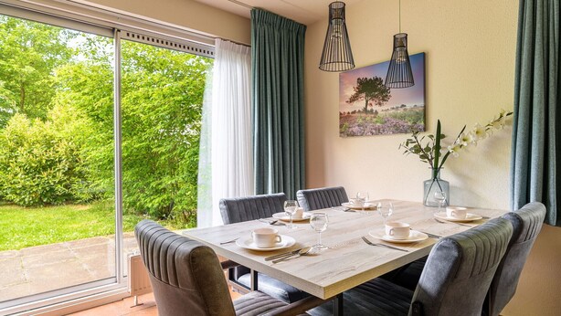 Un comedor con una mesa dispuesta para cuatro personas, un cuadro en la pared y vistas al exterior con vegetación.