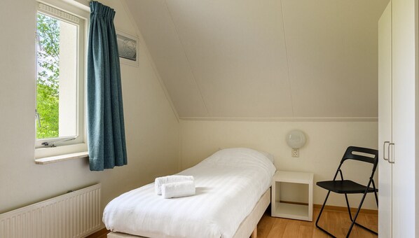 Une petite chambre à coucher, bien rangée, avec un lit simple, une fenêtre avec des rideaux, une table de chevet avec une lampe et une chaise.