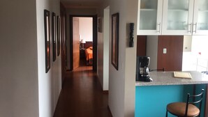 A narrow hallway leading to a bedroom with a bed and a nightstand. A kitchen area with a coffee maker and a chair is visible to the right. - Lovely Suite w/good internet perfect for digital nomads (Quito)