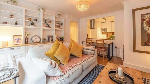 Un salón acogedor con un sofá blanco, cojines amarillos, una mesa de centro de madera y una estantería llena de objetos decorativos.