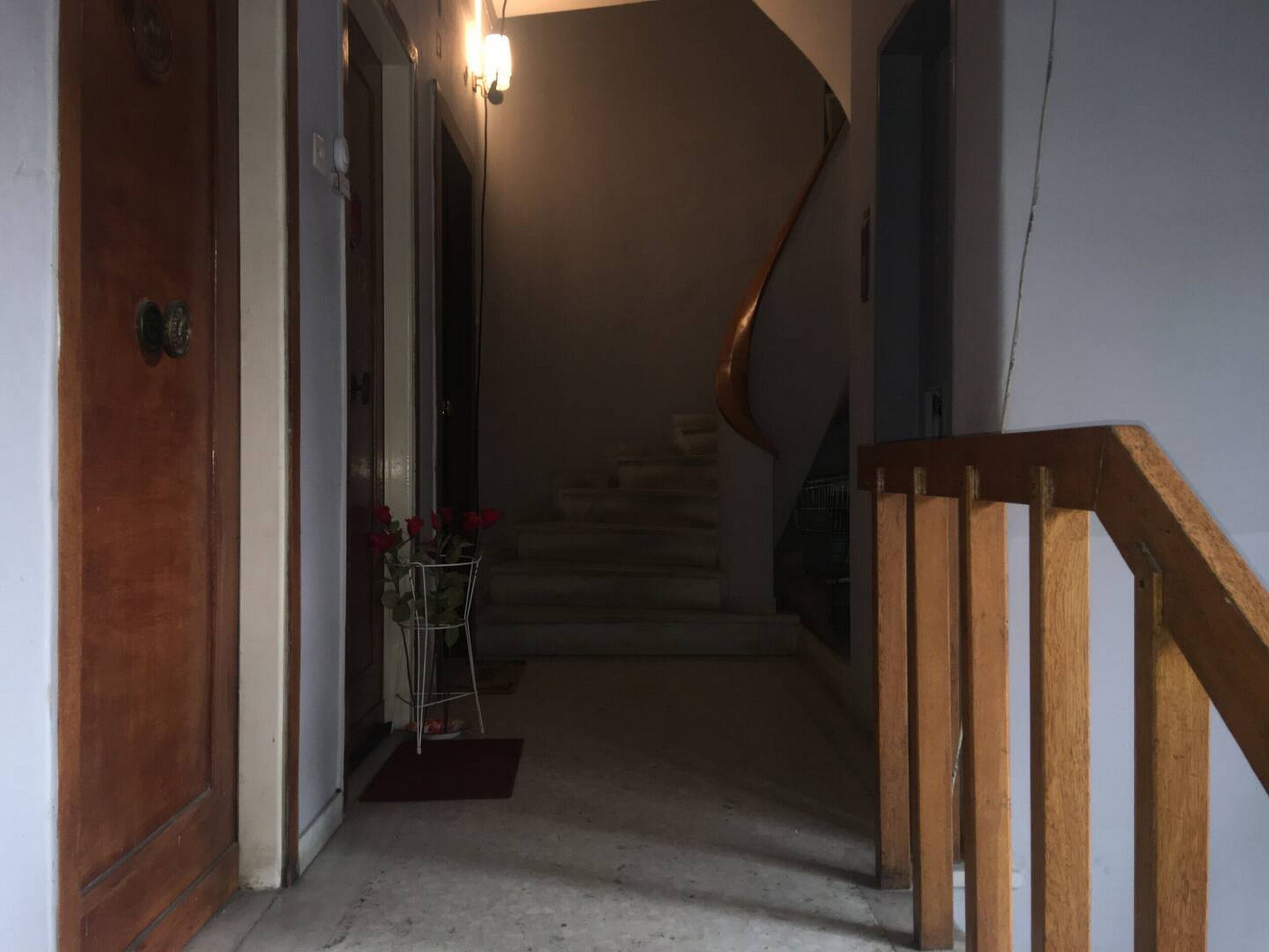 A narrow hallway with a wooden door on the left, a staircase with a wooden railing on the right, and a wall-mounted light fixture above the stairs.