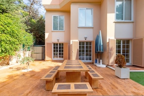 Un patio con una mesa y bancos de madera, rodeado por una casa con ventanas amplias y un árbol pequeño.