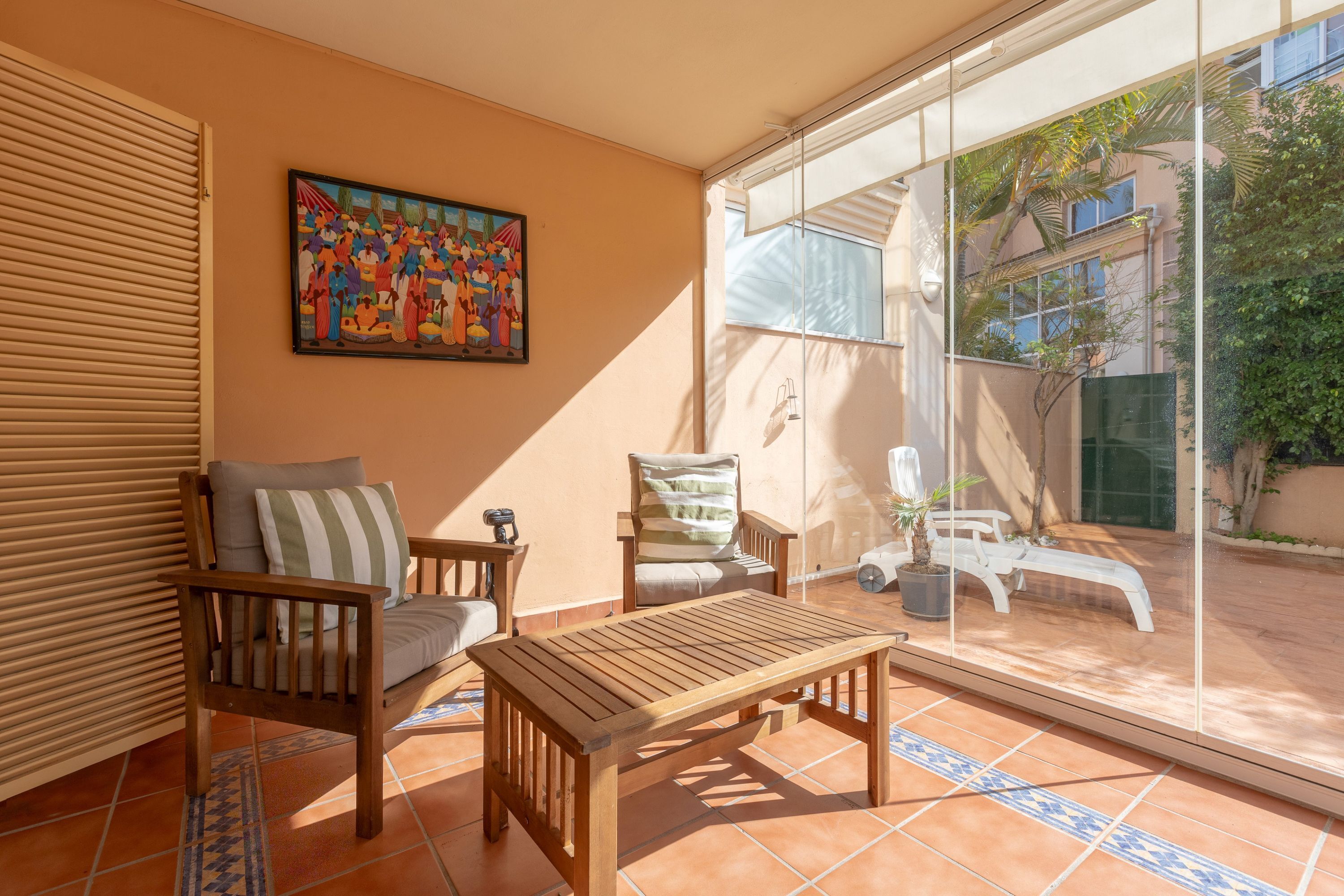 A sunlit patio with a wooden table, two chairs, and a colorful painting on the wall.